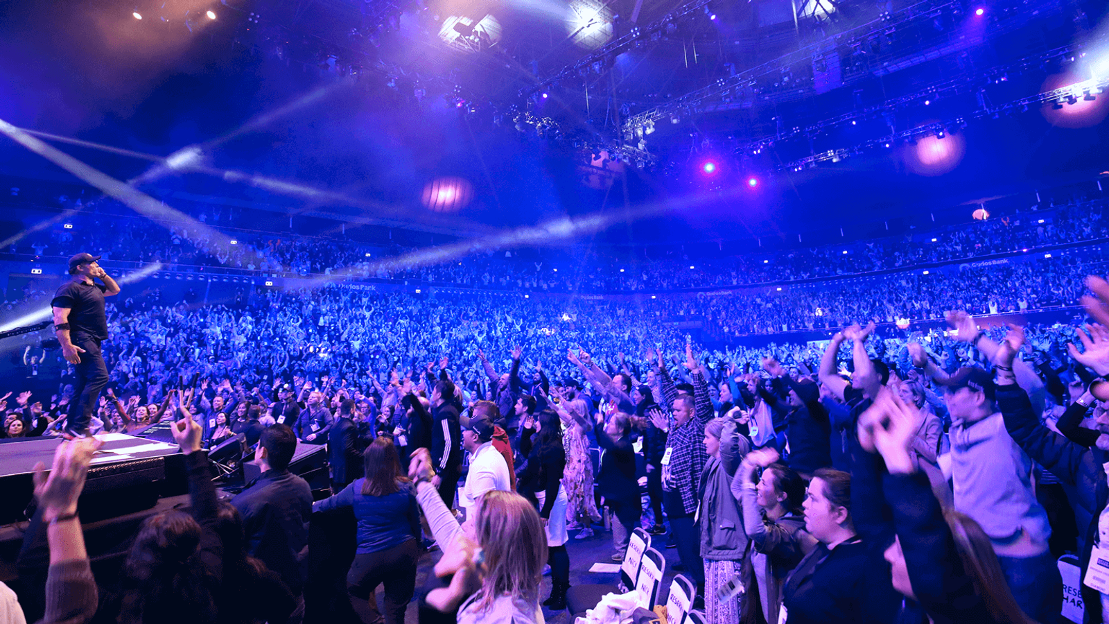 A packed arena crowd with hands raised toward a speaker on stage
