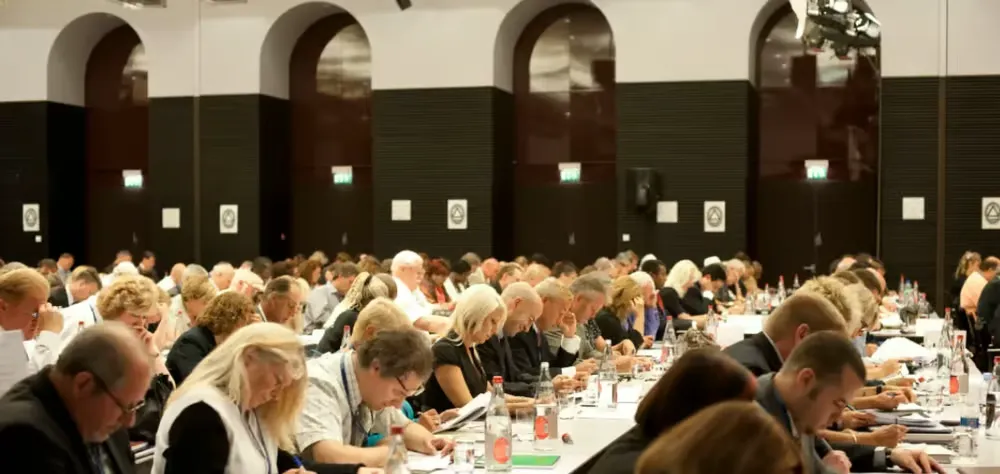 A closed-door assembly of members seated at long tables in a private hall